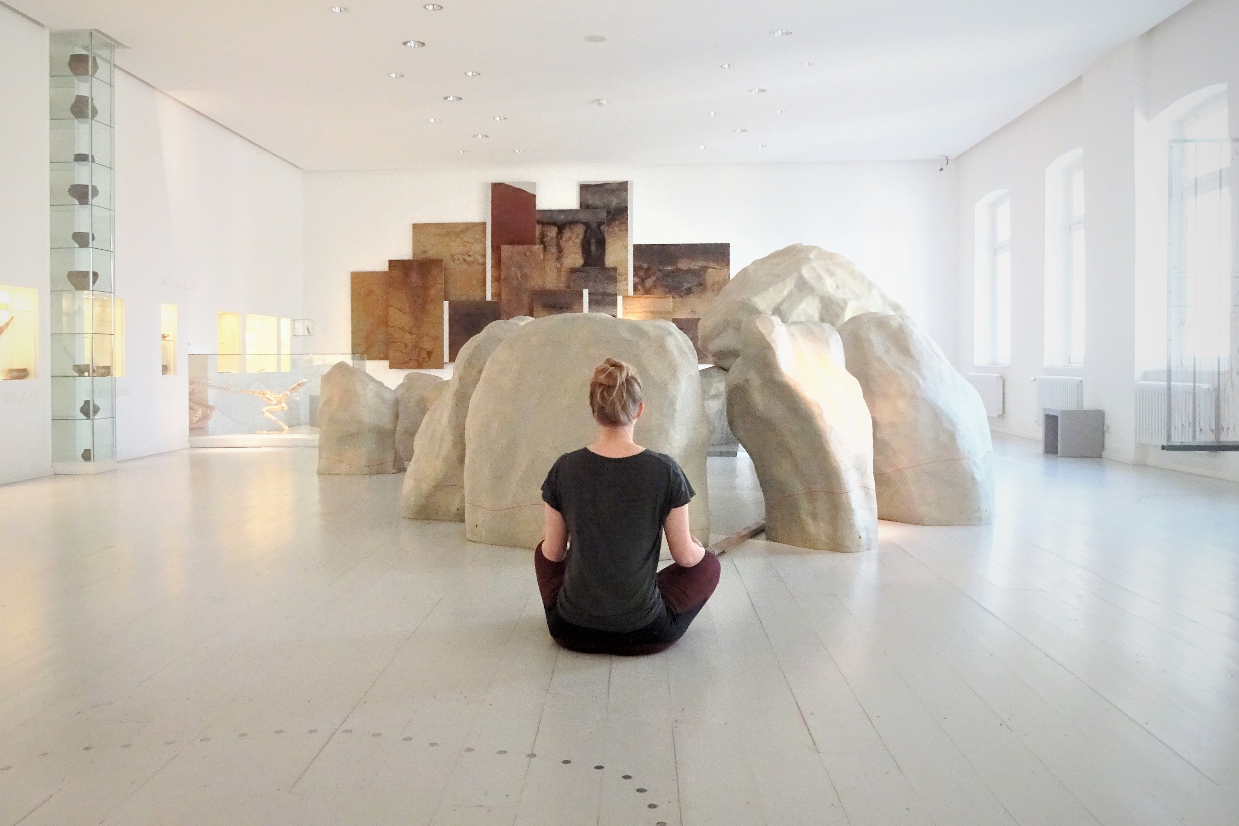 Eine Frau sitzt mit dem Rücken zur Kamera in einem Ausstellungsraum mit großen, unechten Steinen. Sie sitzt im Schneidersitz und es wirkt, als würde sie meditieren.