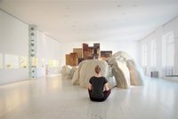 Eine Frau sitzt mit dem Rücken zur Kamera in einem Ausstellungsraum mit großen, unechten Steinen. Sie sitzt im Schneidersitz und es wirkt, als würde sie meditieren.