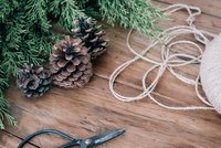 Utensilien zum weihnachtslichen Basteln auf einem Tisch: Ein Tannenzapfen, eine Schere, grüne Tannenzweige und Band.